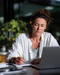 © Milos - A concentrated woman writes in her notebook while working on her laptop in a brightly lit home office, embodying focus and creativity in her workspace.