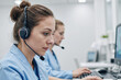 © AnnaStills - Caucasian young adult women working at computers wearing headsets in modern office environment, focusing on screens and performing customer support or call center tasks