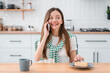 © InsideCreativeHouse - Young woman smiling and talking on a mobile phone, sitting at a kitchen table with a dessert cheesecake and cup of hot coffee tea, relaxing and communicating during a casual morning