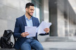 © Prostock-studio - Excited young arab man in formal outfit sitting on the street next to business building, holding documents or CV, having backpack by him, getting ready for job interview, panorama with copy space