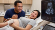 © Николай Срибяник - Pregnant Asian woman and Mixed-Race partner smiling at ultrasound monitor during medical exam in clinic.