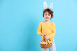© New Africa - Easter celebration. Smiling little boy in headband with bunny ears and wicker basket full of painted eggs on light blue background. Space for text