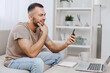 © SHOTPRIME STUDIO - Smiling man using smartphone while sitting on a couch, wearing casual clothes, relaxed atmosphere, modern interior design, engaging with a digital device.