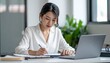 © ChaerunissaH - Focused Asian Woman in White Blouse Writing at Desk with Laptop in Bright Office Space