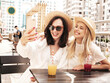 © halayalex - Two young beautiful smiling hipster female in trendy summer white dress and straw hats. Sexy carefree women holding and drinking fresh vegetable cocktail smoothie drink in plastic cup with straw