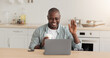 © Prostock-studio - A Black man sits at a kitchen table smiling and waving while on a video call. He holds a cup of coffee in one hand and has a laptop in front of him. Bright lighting fills the kitchen space.
