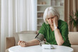 © Anastasiya - Senior caucasian woman sitting with blood pressure monitor on arm at home, elderly female checking hypertension, healthcare and preventive aging concept indoors