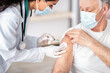 © Prostock-studio - A healthcare worker is giving a vaccine to an older man in a clinic. The man is receiving the shot on his arm while wearing a mask.