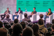 © Anton Gvozdikov - Panel discussion at conference with blurred audience and speakers on stage in bright green backdrop.