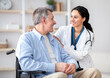© Prostock-studio - A healthcare professional smiles and talks to an older man sitting in a wheelchair. They are in a well-lit room with simple decor. The mood is friendly and supportive, focusing on the conversation.