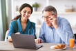© Prostock-studio - A healthcare worker with long hair smiles while helping a senior man with glasses use a laptop. A plate with pastries and a glass of juice is on the table in a well-lit room.