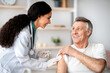 © Prostock-studio - A health professional prepares to give a vaccine to an older man in a clinic. Both are smiling, creating a positive atmosphere. The room has shelves with plants and medical supplies.