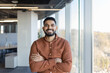© Liubomir - Young professional indian man smiling with crossed arms, standing confidently in a modern office with large bright windows, embodying success, leadership, and positive business attitude