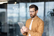 © Liubomir - Young man wearing optical glasses and beard standing in a contemporary office setting, intently looking at a silver tablet, representing digital communication and mobile business