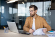 © Liubomir - Young man in glasses focusing on financial data and reports, analyzing numbers and managing business tasks at a modern office desk, utilizing a laptop and calculator for efficiency and organization