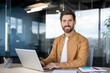 © Liubomir - Smiling man enjoying a productive day, typing on a laptop at a contemporary desk in a bright office environment, embodying professionalism and successful business communication
