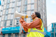 © Iryna - Woman builder wearing safety jacket directs workers on construction site using megaphone overseeing activities