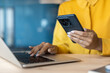 © Tetiana - Close-up photo of the hands of a young woman in a yellow shirt working on a laptop and using a mobile phone in the office