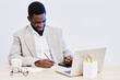 © SHOTPRIME STUDIO - Professional black man in beige blazer and white shirt working at office desk, writing in notebook with laptop, glasses, coffee mug and supplies, modern workspace, business and productivity concept.