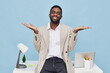 © SHOTPRIME STUDIO - Confident young AfricanAmerican man with beard smiling, wearing casual beige blazer and white shirt, gesturing with open hands, standing in bright office environment with laptop, desk accessories