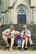 © DSMT - Family sits on steps of church and uses mobile phones while child explores surroundings during sunny day