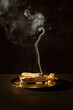 © Simplementbeau. - Bâtons de Palo Santo posés sur un plateau en bronze, un bâton allumé avec une fumée blanche qui s'élève vers le haut finissant en volute, fond brun foncé, ambiance méditative et relaxante