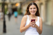 © JackF - Smiling curious young girl around 20 years old stopping briefly while exploring sunlit city streets, using smartphone to take quick travel photo of surroundings