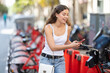 © JackF - Smiling interested young brunette woman using mobile app to unlock rental bike at city station, planning to ride for errands or leisure on summer day