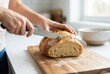 © Stitch - Hands cut a loaf of fresh bread on a wooden board in a kitchen during daylight hours, showing kitchen tools and a simple setup for home cooking