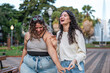 © Nexa - Two diverse female friends with curly hair laughing while walking in a city park during the day.