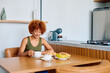 © we.bond.creations - Young woman with curly hair sitting at kitchen table, drinking coffee and having a healthy breakfast with papaya and cheese