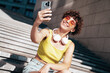 © halayalex - Young beautiful smiling hipster woman in trendy summer clothes. Carefree woman with curls hairstyle, in the street. Positive model holds mobile phone, looks at cellphone screen, uses smartphone apps