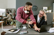 © AnnaStills - Young adult man standing and explaining information to middle aged Caucasian man sitting at desk using laptop, both collaborating in modern office environment