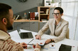 © AnnaStills - Caucasian middle aged woman wearing glasses interviewing young adult man in office, holding resume and gesturing toward document, laptop and paperwork visible on desk
