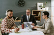 © AnnaStills - Caucasian middle aged man in suit sitting at desk with documents, young adult man with beard and headphones using smartphone, middle aged woman typing on laptop