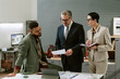 © AnnaStills - Middle aged Caucasian man in suit discussing documents with young adult man holding coffee cup and Caucasian woman taking notes in modern office setting