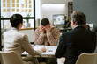 © AnnaStills - Young adult man sitting at desk covering face with hands while two middle aged professionals interviewing him in modern office setting, resumes on table