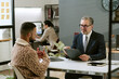 © AnnaStills - Middle aged Caucasian man wearing glasses interviewing young adult man in modern office, both sitting at desk with laptop and clipboard, colleague working in background