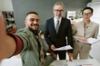 © AnnaStills - Portrait of young adult Middle Eastern man taking selfie with middle aged Caucasian man and middle aged Caucasian woman standing together in modern office