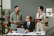 © AnnaStills - Middle aged Caucasian man sitting at desk using laptop while young adult man holding tablet and woman holding notebook standing and discussing business