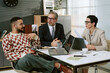 © AnnaStills - Caucasian middle aged man listening to young adult man with beard gesturing while sitting at desk with Caucasian middle aged woman holding documents