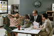 © AnnaStills - Caucasian middle aged man in suit discussing business strategy with young adult man and woman in modern office, all sitting at table with laptops and documents