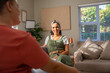 © wavebreak3 - Diverse female friends chatting on sofa in living room, wearing coral t-shirt, olive jumpsuit