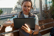 © shustrilka - Attractive caucasian woman sitting in park using tablet for remote work, teleconference concept