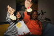 © Studio Marmellata - Two young women share a bag of potato chips while relaxing on a couch, offering a chip to the camera.