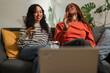 © Studio Marmellata - Two young women laugh while watching something on a laptop and eating snacks on a couch.