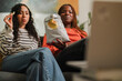 © Studio Marmellata - Two women relax on a couch, sharing a bag of chips and watching a screen.