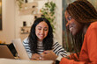 © Studio Marmellata - Two young women are smiling and interacting while working on laptops in a bright, modern cafe setting.