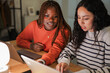 © Studio Marmellata - Two young women collaborate on a project using laptops at a desk, illuminated by a desk lamp.