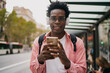 © BullRun - Urban Black man smiling directly at camera with headphones on and smartphone in hand, radiating confidence and digital ease while waiting at city bus stop